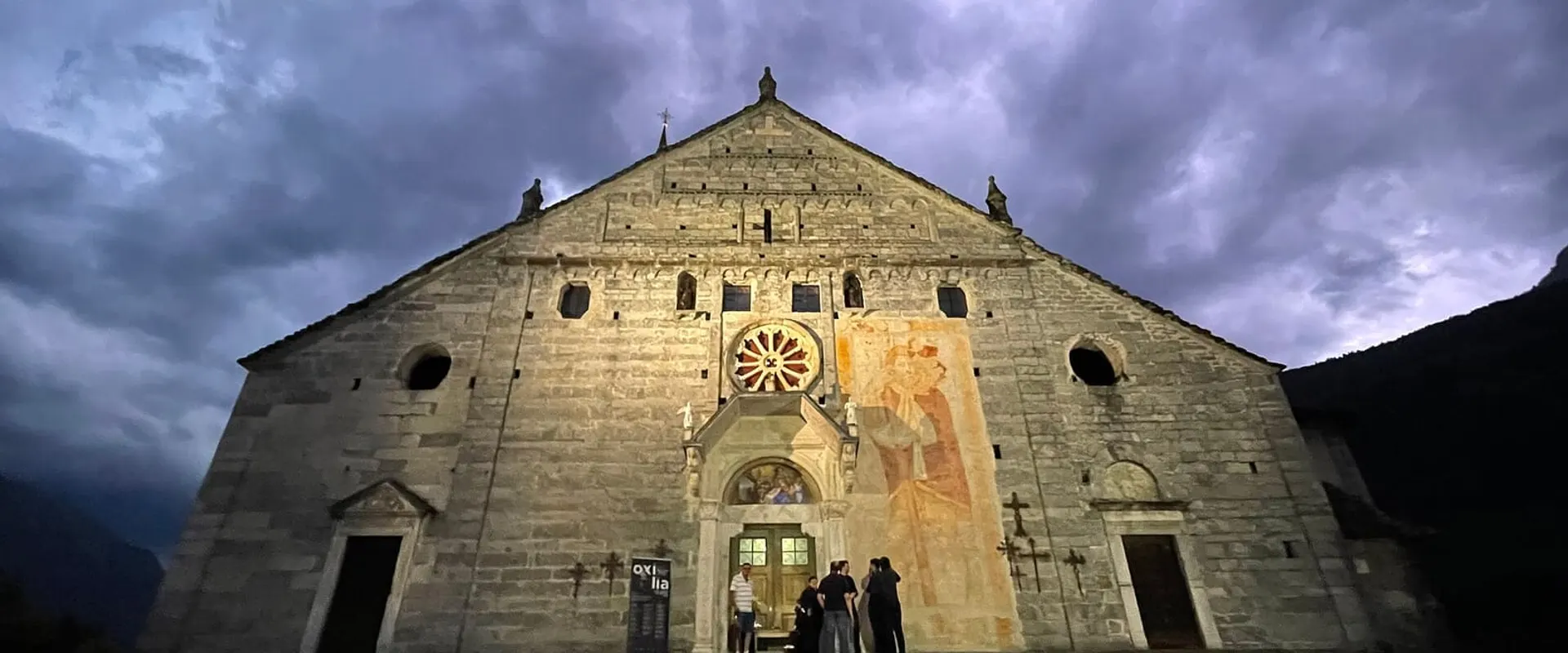 Facciata in pietra della chiesa con il rosone e un grande affresco, al tramonto