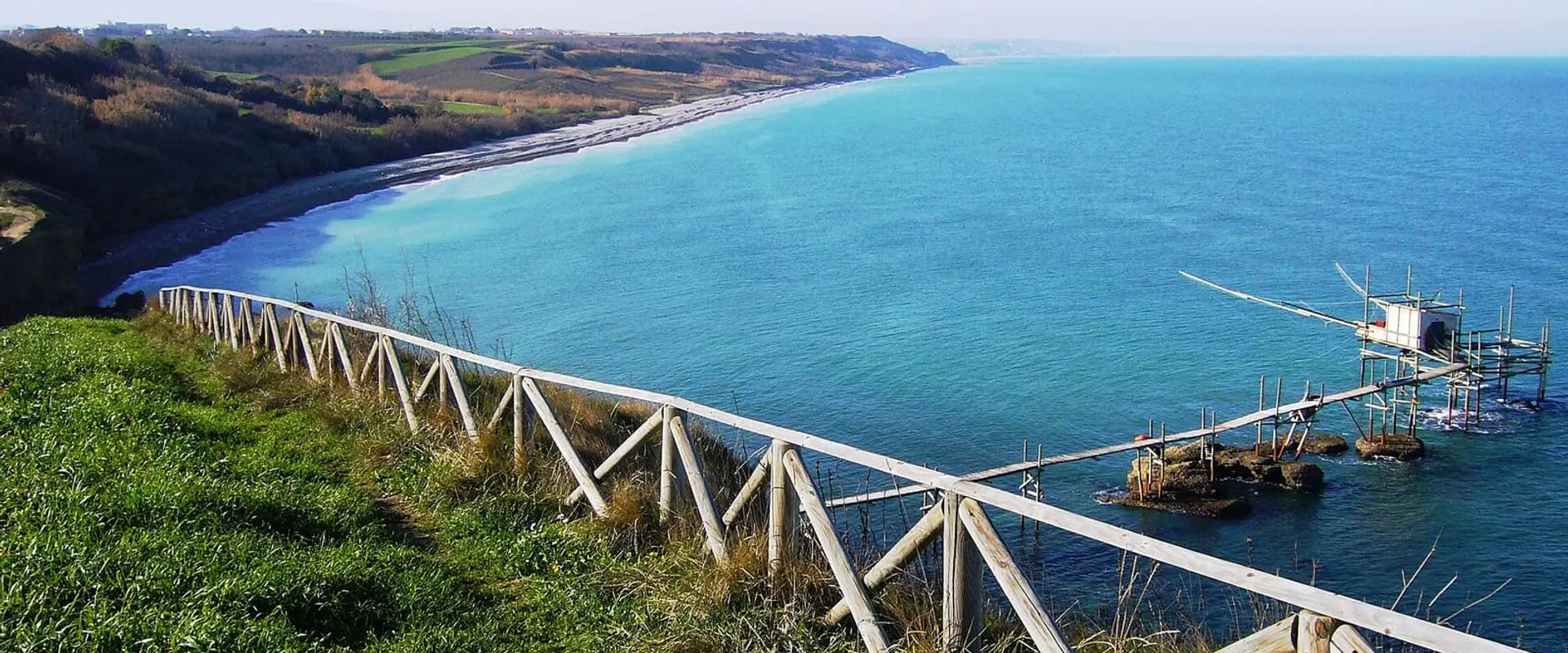Vista dall’alto della costa con il mare azzurro, la staccionata del belvedere e un trabocco in basso