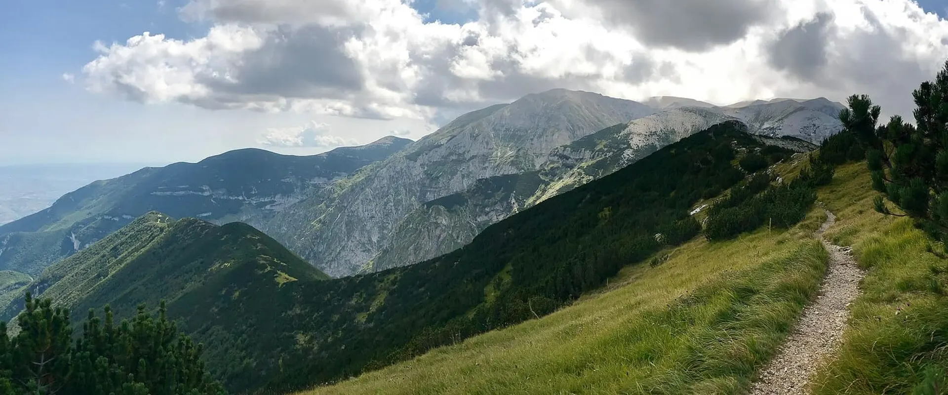 Sentiero che sale lungo un crinale erboso verso le montagne, sotto un cielo con le nuvole