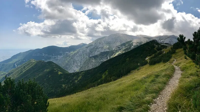 Sentiero che sale lungo un crinale erboso verso le montagne, sotto un cielo con le nuvole